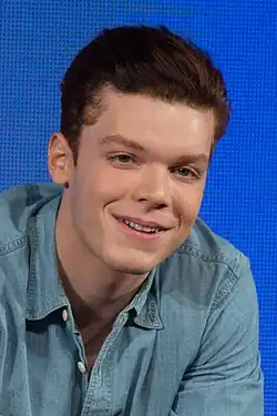 A 29-year-old man with long, brown hair, smiling at someone to the right of the camera.