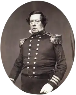 Black and white photo of a clean-shaven, unsmiling man with bags under his eyes. He wears a dark uniform with a double row of buttons and epaulettes.