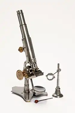 A stand microscope created by Joseph Gutteridge, a weaver from Coventry. The microscope allows coarse and fine adjustments, and its height is adjustable by a set screw. It consists of two objectives each with a screw on cap and a slidecarrier that is moveable laterally. The mirror also has a cap. It has a bench magnifier, lens moveable through 360° and has a base weighted with lead.
