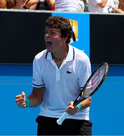 Raonic with his mouth open looks to the left, presumably to the crowd, and holds his racquet in his left hand.