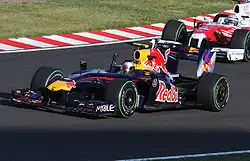 a dark blue F1 car drives along a track ahead of a pink F1 car