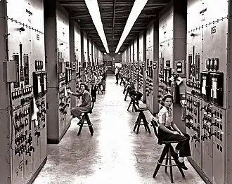 A long, high hallway, with bulky calutrons covered in switches and dials rising from the floor to the ceiling. Women sit on high chairs operating the machinery.